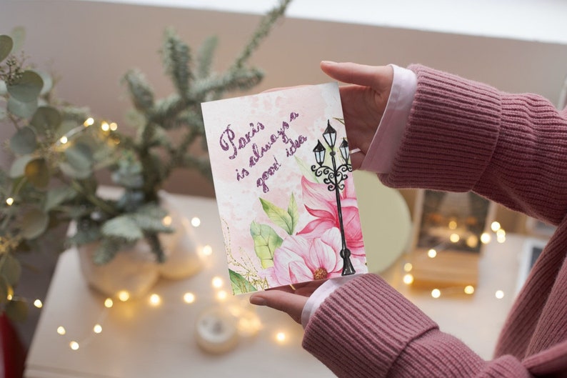 Pu&ograve; includere: Un biglietto di auguri con la scritta "Paris is always a good idea" in corsivo viola. Il biglietto presenta un'illustrazione ad acquerello di un lampione e fiori rosa. Il biglietto &egrave; tenuto da una persona che indossa un maglione rosa.