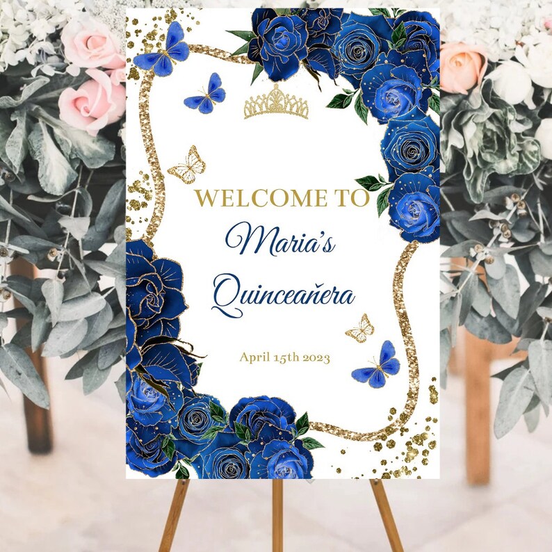 May include: A white welcome sign with gold glitter and blue roses. The sign reads "Welcome to Maria's Quincea&ntilde;era April 15th 2023".