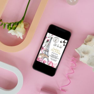 Pu&ograve; includere: Uno smartphone nero mostra un invito di compleanno con la Torre Eiffel, fiori di magnolia e farfalle. L'invito dice "Oh la la! 21&deg; compleanno di Charlotte". Il telefono &egrave; su una superficie rosa con elementi decorativi.