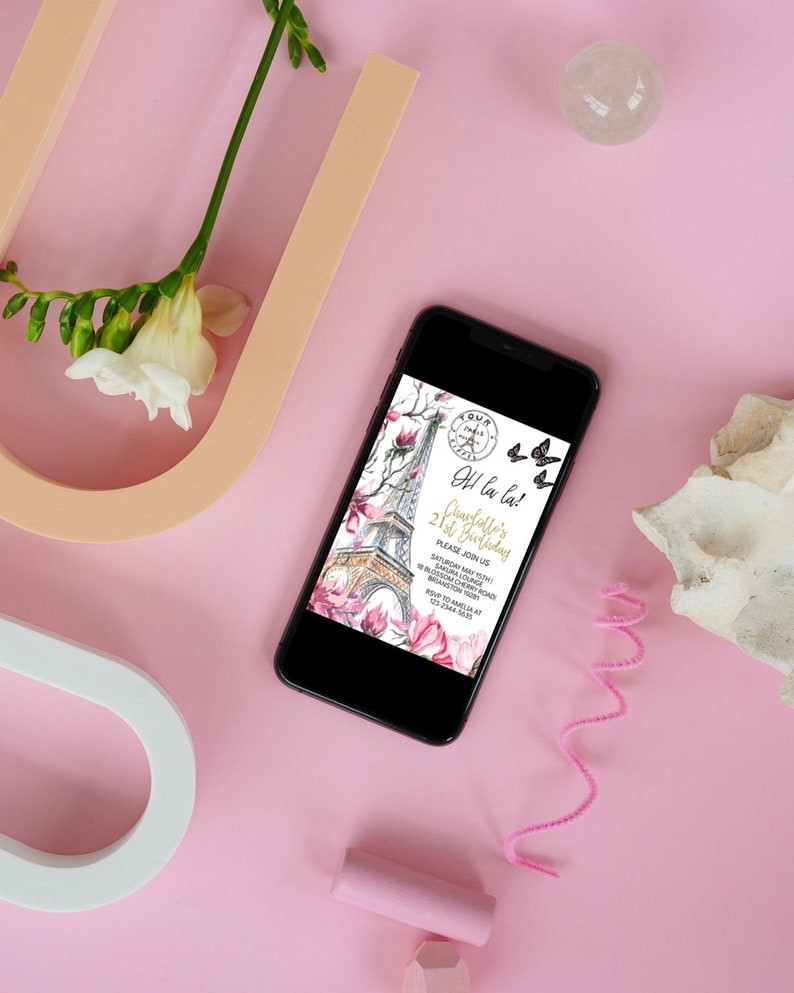 Pu&ograve; includere: Uno smartphone nero mostra un invito di compleanno con la Torre Eiffel, fiori di magnolia e farfalle. L'invito dice "Oh la la! 21&deg; compleanno di Charlotte". Il telefono &egrave; su una superficie rosa con elementi decorativi.