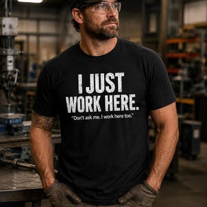 Puede incluir: Una camiseta negra de manga corta con el texto blanco desgastado I JUST WORK HERE. y el texto más pequeño Don't ask me. I work here too. impreso en el pecho. La camiseta tiene cuello redondo y un corte estándar, hecha de algodón.