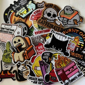 May include: A collection of embroidered patches with various designs and text. The patches feature images of cats, a skull, a slice of toast reading a book, and phrases like "Dead Inside" and "Everything's Fine."