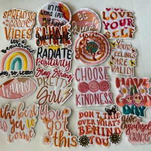 May include: A collection of embroidered patches with inspirational quotes and designs. The patches feature phrases like "Spread Good Vibes," "Love Yourself," and "Choose Kindness." Colors include pink, orange, yellow, and white.