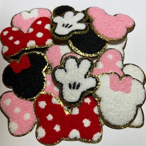 May include: A collection of embroidered patches featuring iconic cartoon character designs. The patches include mouse ears in pink, black, and white, with gold trim. Some patches feature polka dots and bows in red and pink.