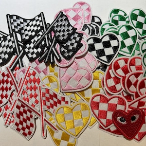 May include: A collection of embroidered patches in various shapes and colors. The patches include checkered flags in black, red, and white, and heart shapes in pink, green, yellow, and red with checkered patterns. The patches are likely for clothing or accessories.