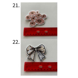 May include: Two embroidered patches: one of a cluster of pink flowers, the other of a white and gold bow. A red ruler is placed next to each patch. The flower patch measures approximately 2 inches in length. The bow patch measures approximately 2.5 inches in length.
