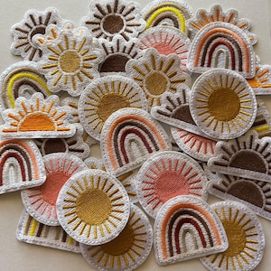 May include: A collection of embroidered patches featuring sun and rainbow designs. The patches are in various shapes and sizes, with a color palette of yellow, orange, brown, and pink. The patches have white borders and are likely for clothing or accessories.