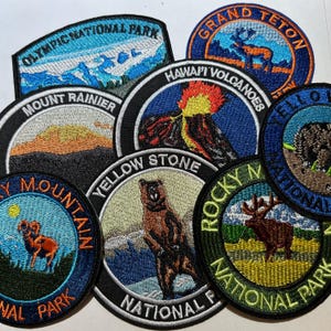 May include: A collection of embroidered patches featuring national park designs. Patches include Olympic, Mount Rainier, Grand Teton, Hawaii Volcanoes, Yellowstone, and Rocky Mountain National Park. Each patch has a unique design and color scheme.