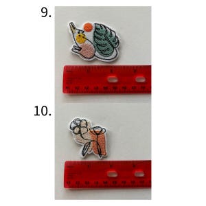 May include: Two embroidered patches. The top patch features a bird with a yellow chest, a red sun, and a green leaf. The bottom patch shows a flower and a vase in beige, orange, and black. Each patch is approximately 1 inch in size.
