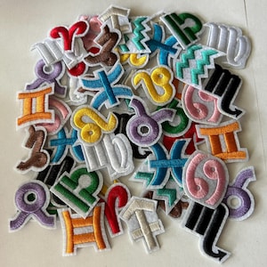 May include: A collection of embroidered patches featuring colorful zodiac symbols. Each patch has a white border and showcases a unique design in various colors, including red, yellow, blue, and green. The patches are scattered across a white surface.