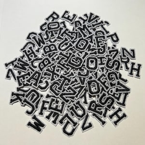 May include: A pile of black embroidered varsity letter patches with white borders. The letters are in a classic block font, perfect for customizing apparel or accessories. The patches are scattered on a white surface.