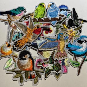 May include: A collection of embroidered bird patches in various colors, including blue, green, orange, and yellow. The patches depict different bird species, such as hummingbirds and bluebirds, with detailed stitching and a white border. These patches are ideal for adding to clothing or accessories.