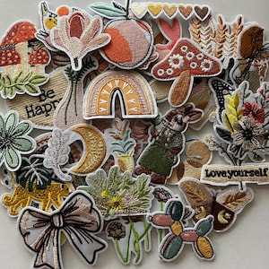 May include: A collection of embroidered patches in various shapes and colors. Designs include a rainbow, mushrooms, flowers, a peach, a bunny, a bow, and text that says "Be Happy" and "Love yourself". The patches have a white border.