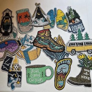 May include: A collection of embroidered patches with outdoor and camping themes. Designs include a hiking boot, tent, mountain range, coffee mug with "Drink Coffee Outdoors", and a yellow van. Patches are in various colors with detailed stitching.