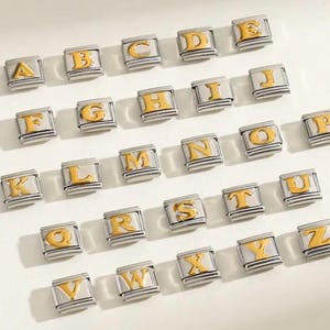 May include: A collection of silver-toned alphabet charms with gold-colored letters. Each charm is a small square with a letter from A to Z. The charms are arranged in rows and columns on a white surface.