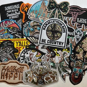May include: A collection of embroidered patches with various designs and text. Patches include a bow, deer, and phrases like "Someone Pour Me Up A Double Shot Of Whiskey" and "Mama's Let Your Boys Be Country". Colors include black, brown, green, and tan.