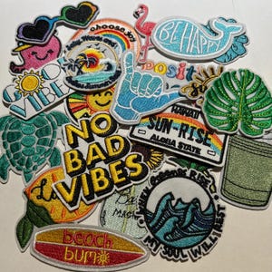 May include: A collection of embroidered patches with various designs and text. The patches include a turtle, a whale that says "Be Happy", a surfboard that says "beach bum", and a patch that says "No Bad Vibes".