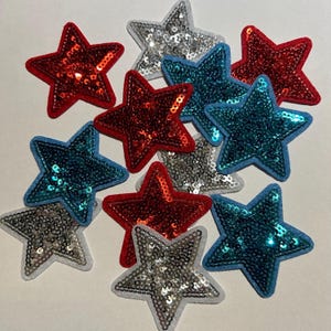 May include: A collection of star-shaped patches in red, silver, and blue. Each star is covered in sequins and has a felt border. The patches are scattered on a white surface, creating a festive and decorative appearance.