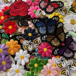 May include: A collection of colorful embroidered patches featuring various floral and insect designs. The patches include red roses, daisies, butterflies, and bees in shades of pink, blue, yellow, and green. These patches are ideal for adding decorative flair to clothing or accessories.