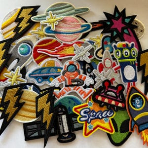 May include: A collection of colorful embroidered patches with space-themed designs. The patches include planets, rockets, astronauts, lightning bolts, and the word "Space" in a starburst. The patches are in various shapes and sizes.
