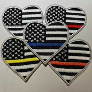 May include: Heart-shaped patches featuring the American flag design. Each patch has a white border and a different colored stripe: red, blue, yellow, and orange. The patches are embroidered with stars and stripes.