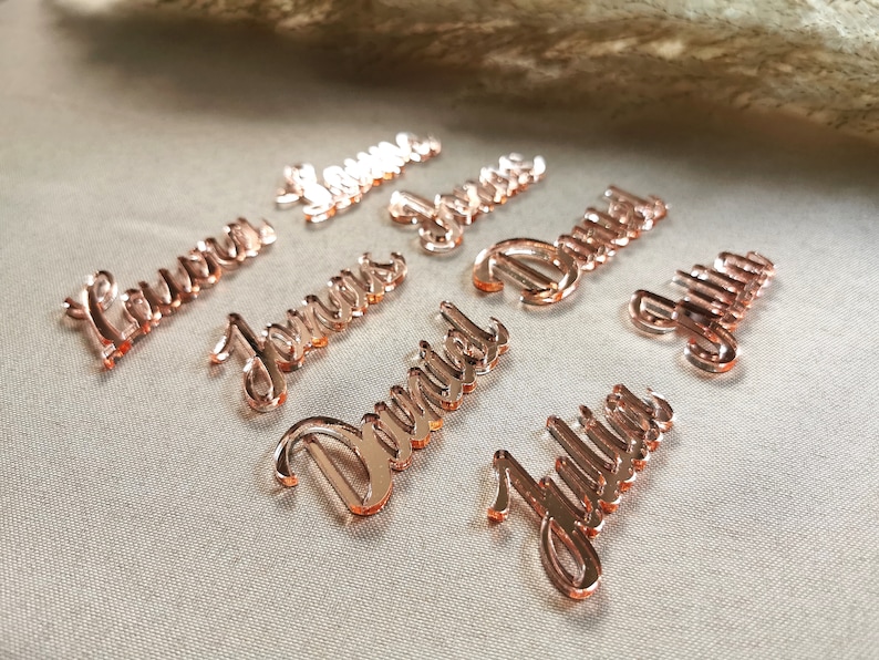 May include: Rose gold acrylic place cards with the names Laura, Jenners, Daniel, Julia, and others. The names are written in a cursive font.