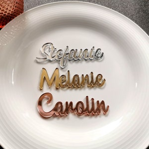 May include: Three acrylic place cards with names written in cursive. The names are Stephanie, Melanie, and Carolin. The place cards are silver, gold, and rose gold.