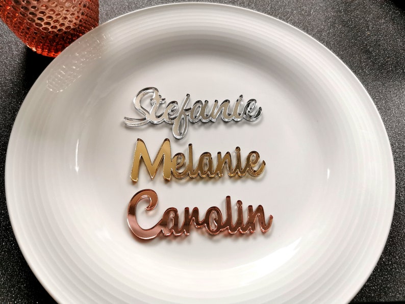May include: Three acrylic place cards with names written in cursive. The names are Stephanie, Melanie, and Carolin. The place cards are silver, gold, and rose gold.