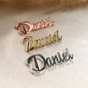 May include: Three acrylic nameplates with the name "Daniel" written in a cursive font. The nameplates are in three different colors: rose gold, gold, and silver.