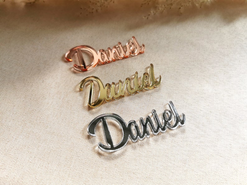 May include: Three acrylic nameplates with the name "Daniel" written in a cursive font. The nameplates are in three different colors: rose gold, gold, and silver.