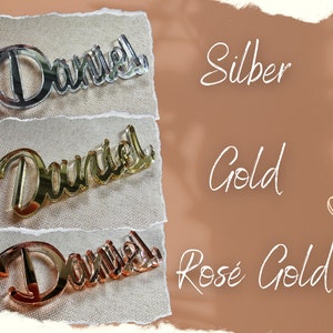 May include: Three mirrored acrylic name plaques with the name "Daniel" in a cursive font. The plaques are displayed on a beige fabric background. The plaques are shown in three different colors: silver, gold, and rose gold.