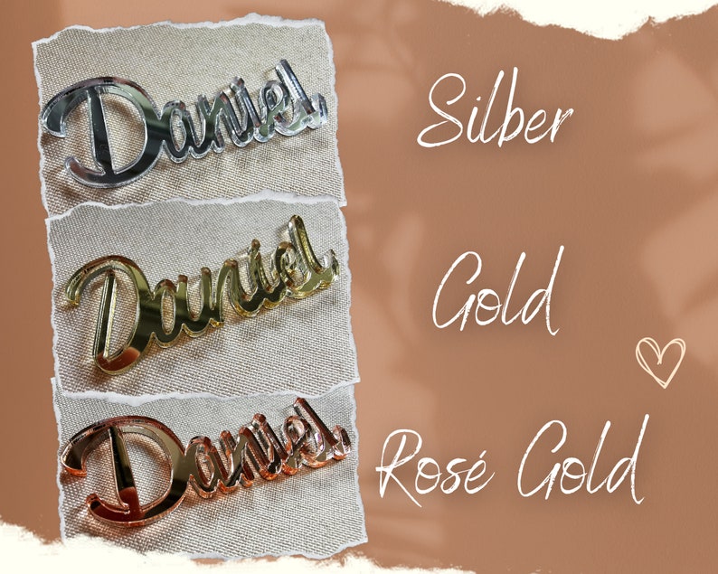 May include: Three mirrored acrylic name plaques with the name "Daniel" in a cursive font. The plaques are displayed on a beige fabric background. The plaques are shown in three different colors: silver, gold, and rose gold.