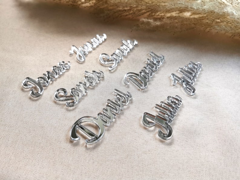 May include: Silver mirror acrylic place cards with the names "Jenna", "Sarah", "Daniel", "Julia", "James", "David", and "Justin" written in a cursive font.