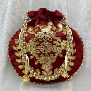 May include: A maroon drawstring bag with gold embroidery and a white pearl beaded trim. The bag has two tassels with gold bells.