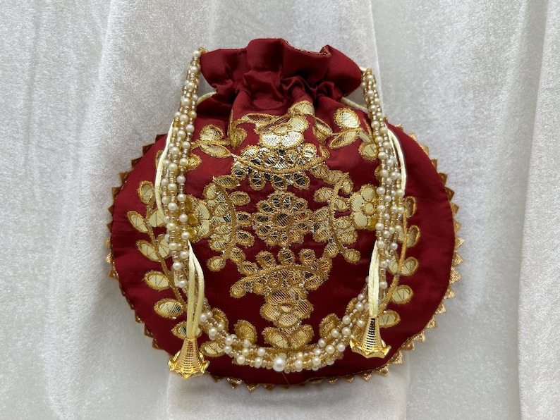 May include: A maroon drawstring bag with gold embroidery and a white pearl beaded trim. The bag has two tassels with gold bells.