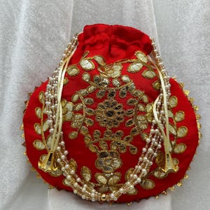 May include: A red drawstring bag with gold embroidery and white pearl beads. The bag has a scalloped edge and is decorated with floral patterns.