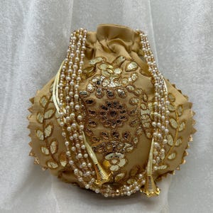 May include: A gold and cream drawstring bag with a floral design and pearl embellishments. The bag has a long, beaded strap.