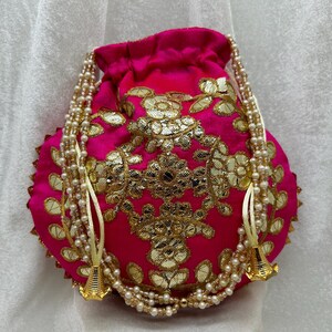 May include: A pink drawstring bag with gold embroidery and pearl accents. The bag has a round shape and is decorated with floral patterns.