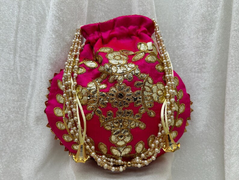 May include: A pink drawstring bag with gold embroidery and pearl accents. The bag has a round shape and is decorated with floral patterns.