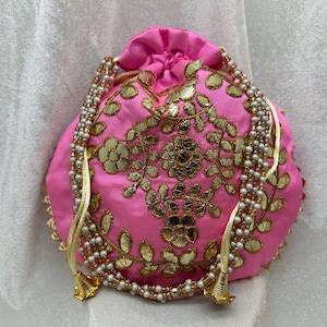 May include: A pink drawstring bag with gold embroidery and pearl accents. The bag has a floral design and is decorated with small, round mirrors.