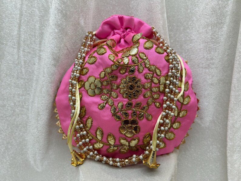 May include: A pink drawstring bag with gold embroidery and pearl accents. The bag has a floral design and is decorated with small, round mirrors.