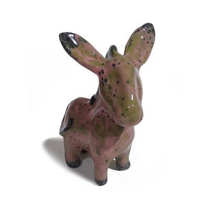 May include: A ceramic donkey figurine in shades of pink, green, and black. The donkey has large ears and a spotted pattern. The figurine is standing on four legs and is set against a white background.