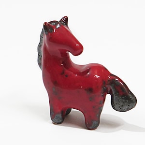 Puede incluir: Una pequeña figura de caballo de cerámica roja con detalles negros. El caballo está de pie sobre sus cuatro patas y tiene una cola larga.