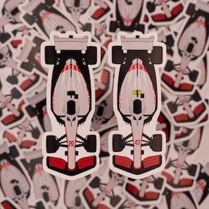 May include: Two white Formula 1 race car stickers with red and black accents. The stickers feature the numbers "20" and "47" on the front of the cars.