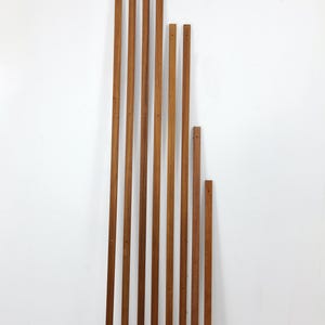 Könnte beinhalten: Eine Ansammlung von Holzlatten unterschiedlicher Länge, die vertikal an einer weißen Wand angeordnet sind. Die Latten sind warmbraun, mit sichtbarer Holzmaserung und kleinen Löchern. Die Anordnung deutet auf ein potenzielles dekoratives oder strukturelles Element hin.