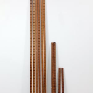 Puede incluir: Un conjunto de listones de madera para somier en varias longitudes. Los listones, de color marrón cálido, presentan agujeros espaciados uniformemente. El conjunto se dispone contra una pared blanca, sugiriendo una estética minimalista.