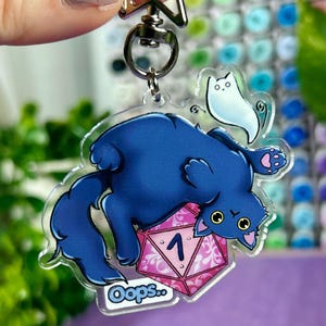 Könnte beinhalten: Ein klarer Acryl-Schlüsselanhänger mit einer blauen Katze und einer weißen geisterhaften Figur. Die Katze liegt auf einem rosa-weißen Würfel mit der Zahl 1. Der Schlüsselanhänger hat einen silbernen sternförmigen Verschluss und das Wort "Oops..." ist auf dem Würfel geschrieben.