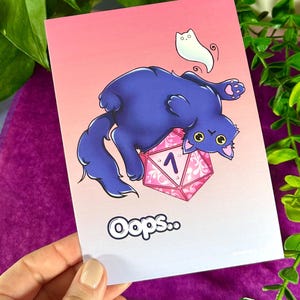 Könnte beinhalten: Eine Grußkarte mit einer Cartoon-Katze und einem D20-Würfel. Die Katze ist dunkelblau und liegt auf dem Rücken, mit einem kleinen weißen Geist darüber. Der Würfel ist rosa und weiß. Das Wort "oops" steht unten.