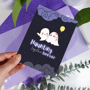 May include: A black card with a cute illustration of two ghosts holding hands. The card has the text "Haunting together forever" in white lettering.