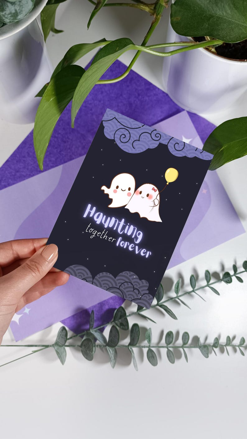 May include: A black card with a cute illustration of two ghosts holding hands. The card has the text "Haunting together forever" in white lettering.